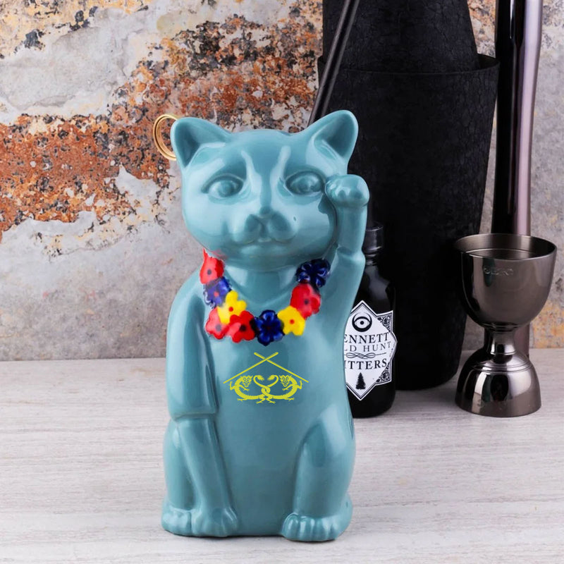 Blue cat-shaped bottle with a colorful lei on a bar counter with a bottle and jigger in the background.