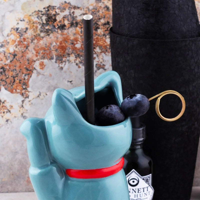 Blue cat-shaped drink mixer with a black straw and blueberries, next to a bottle of Gin and Tonic.